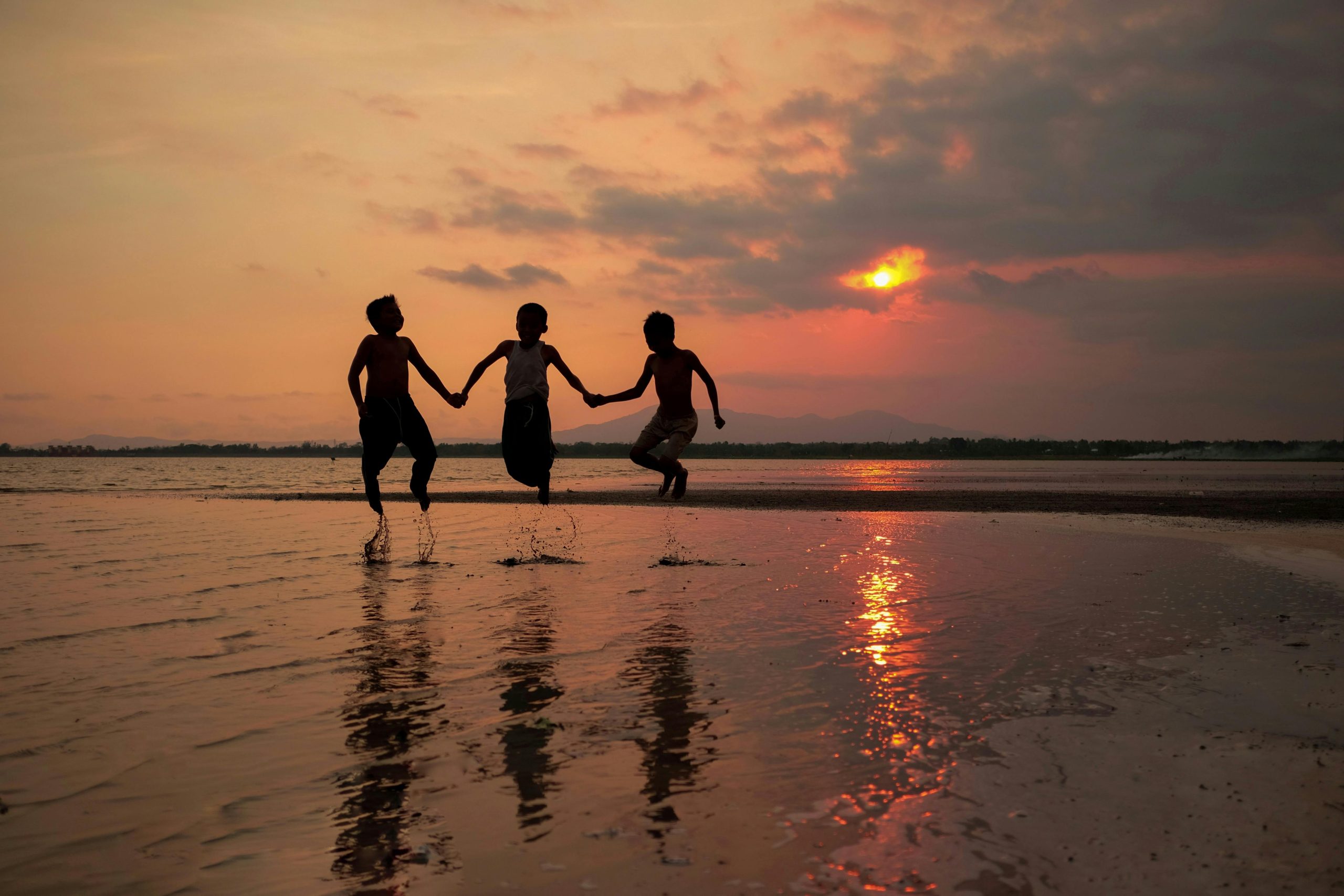 Three children hold hands and jump jubilantly on a sandy beach during a vibrant sunset in Indonesia.