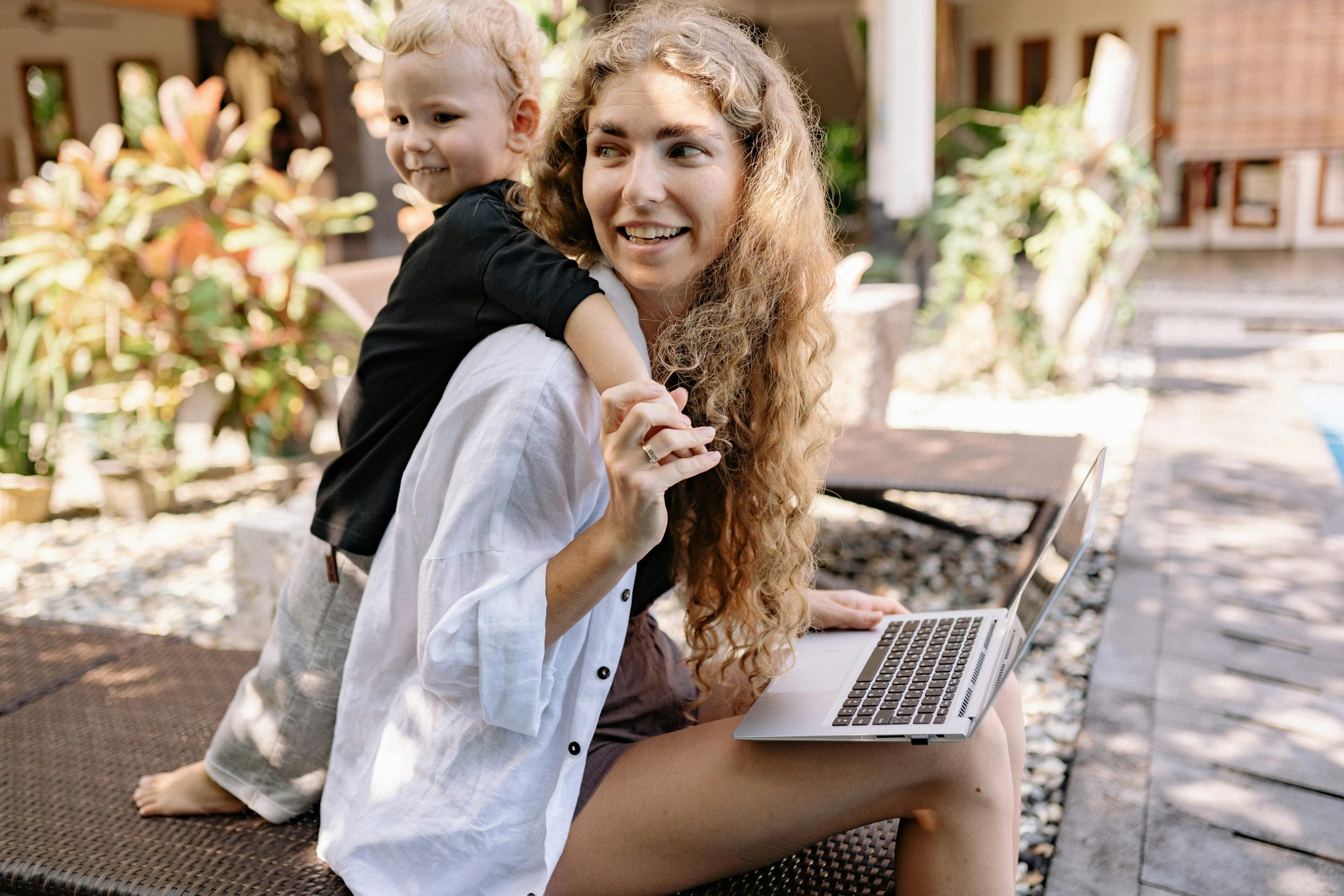 Smiling woman with curly hair holding child in outdoor setting with greenery and laptop.